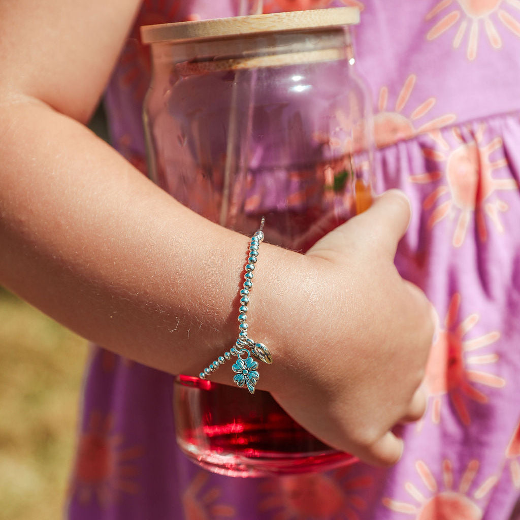 A close-up of a child's arm wearing a silver-toned beaded bracelet with a clover good luck charm, holding a glass jar with a wooden lid and red liquid, wearing a purple sun-patterned dress.