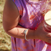 A close-up outdoor shot of a child wearing a silver beaded bracelet with a small butterfly charm, holding a glass jar. The child is wearing a purple dress with orange sun patterns.