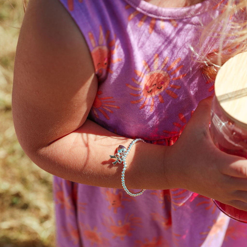 A close-up outdoor shot of a child wearing a silver beaded bracelet with a small butterfly charm, holding a glass jar. The child is wearing a purple dress with orange sun patterns.