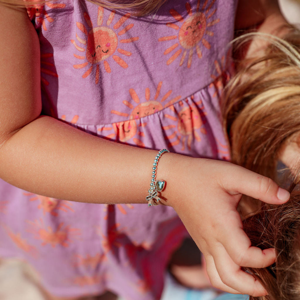 A close-up lifestyle shot of a child wearing a delicate silver beaded bracelet with a small charm shaped like a bow, while another child in a purple sun-print dress gently touches their hair.