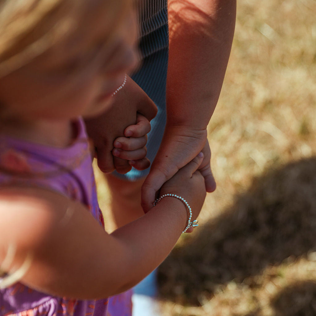 A close-up outdoor photo of a young child in a purple floral dress holding another child's hand. The child is wearing a delicate silver beaded bracelet on one wrist.