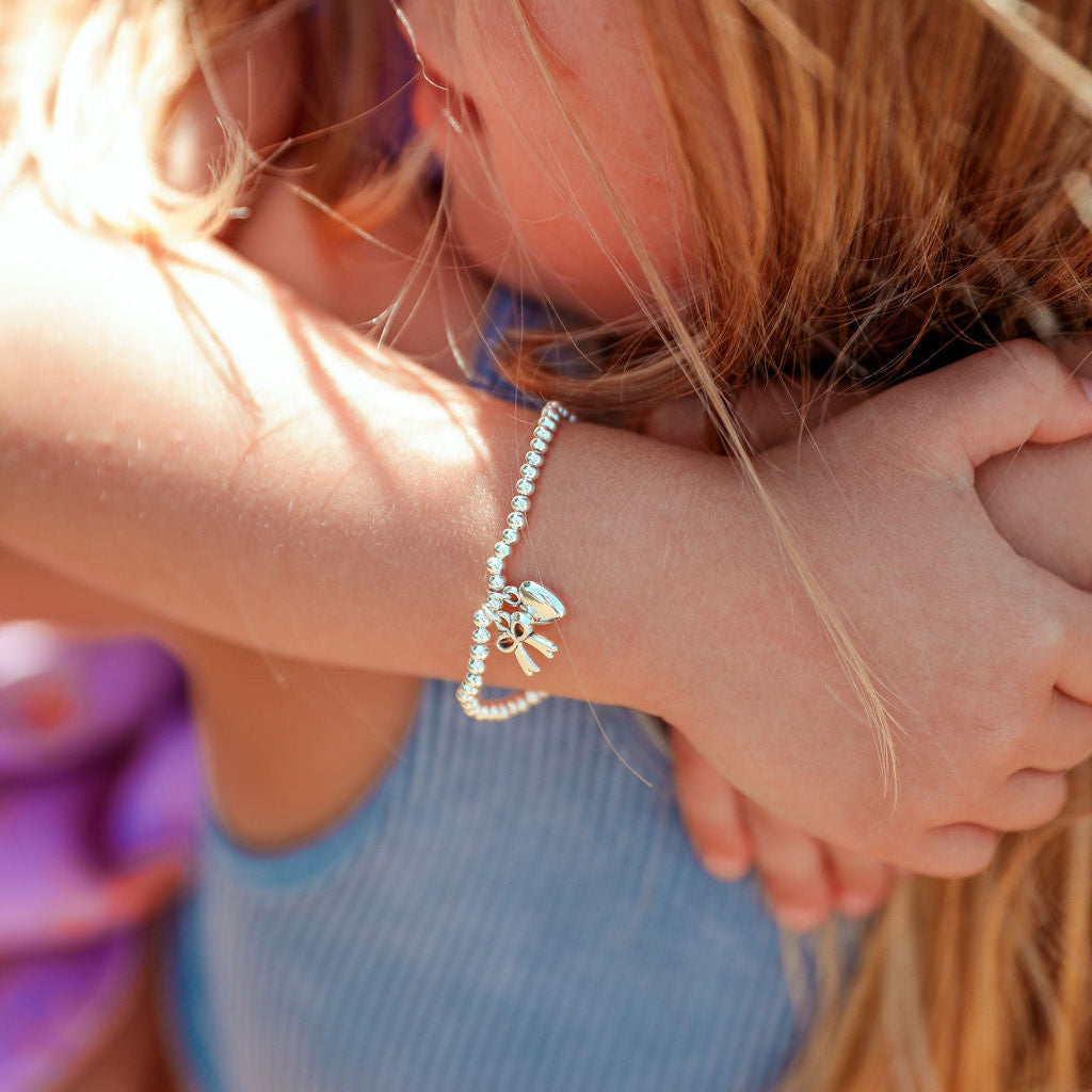 A close-up lifestyle shot of a child embracing another child wearing a delicate silver beaded bracelet with a small flower charm on her wrist, paired with a light blue ribbed crop top.