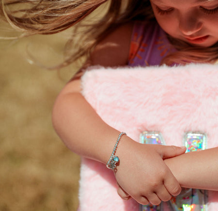 A close-up lifestyle shot of a young girl wearing a silver chain bracelet with a blue-green stone, resting her arm on a fluffy pink blanket with holographic letter 'N' notebook.
