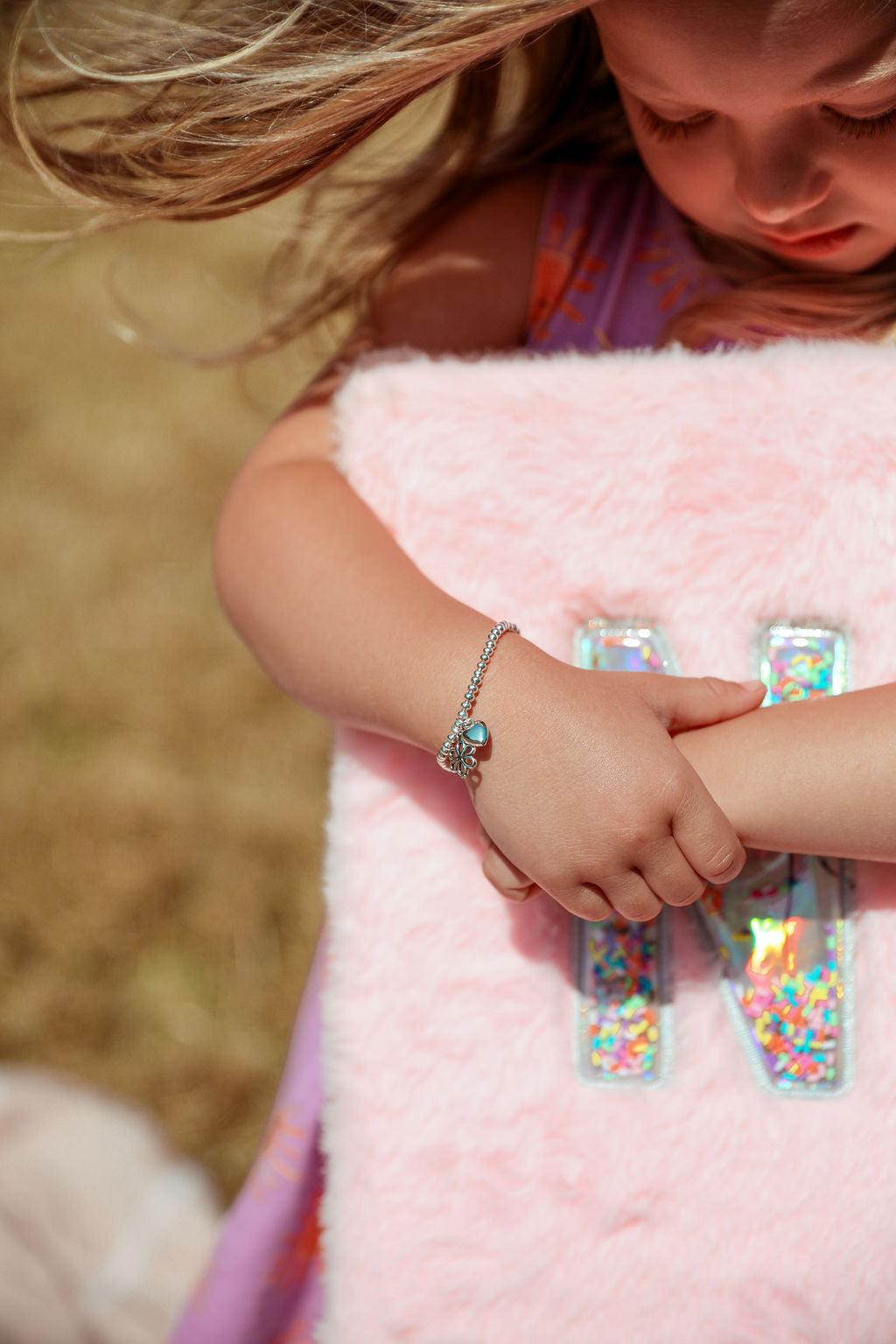 A close-up lifestyle shot of a young girl wearing a silver beaded bracelet with a blue-green stone, resting her arm on a fluffy pink notebook with holographic letter 'N'.