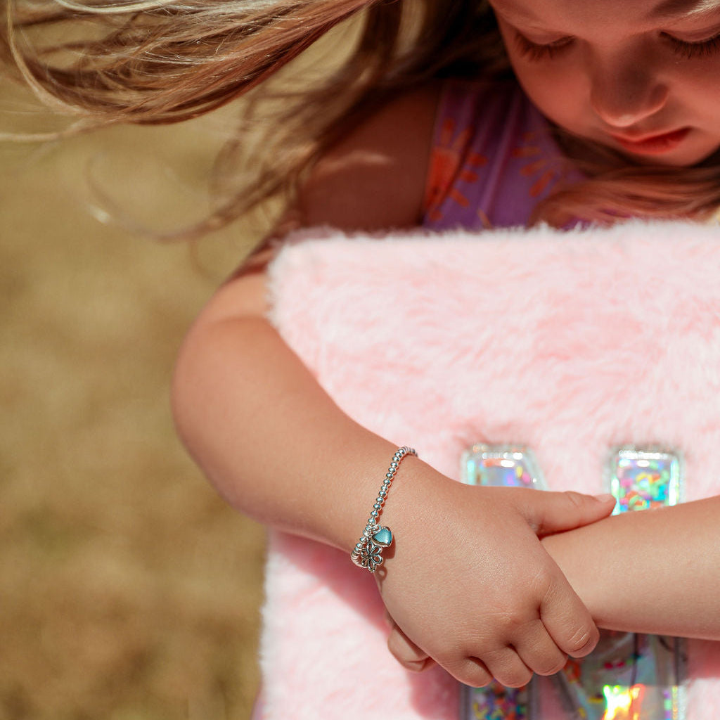 A close-up lifestyle shot of a young girl wearing a silver chain bracelet with a blue-green stone, resting her arm on a fluffy pink blanket with holographic letter 'N' notebook.