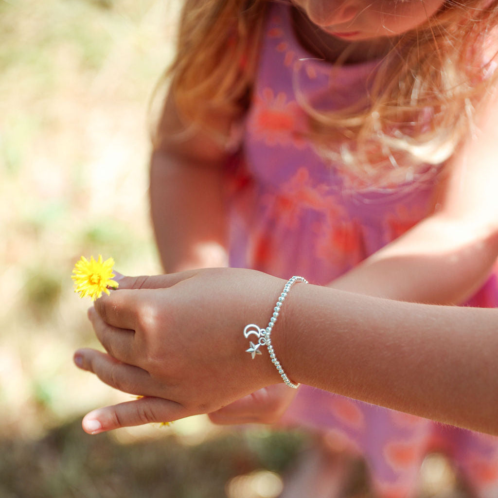 A close-up of a child's hands holding a small yellow flower outdoors, wearing a delicate silver beaded bracelet with a bow charm. The child is wearing a pink patterned dress, and the background is a soft-focus natural outdoor setting.