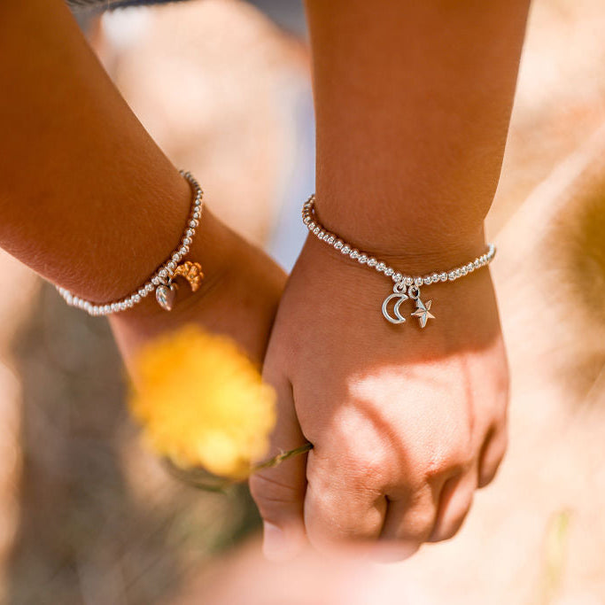 Two close-up views of hands holding and touching delicate silver charm bracelets, one with a crescent moon and the other with a star charm, set against a warm, blurred outdoor background.