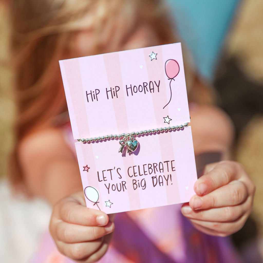 A child holds up a pink card featuring a beaded bracelet with small charms. The card reads 'HIP HIP HOORAY' and 'LET'S CELEBRATE YOUR BIG DAY!' with balloon and star illustrations.