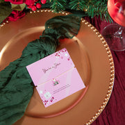 A heartfelt christmas gift for her with a heart and star charm bracelet, displayed on a gold charger plate with a dark green napkin. The setting is decorated with red roses, green pine branches, and festive ornaments.