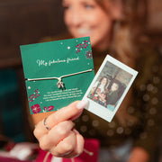 A woman holds up a heartfelt bracelet gift on a green card next to a printed photo, with a blurred festive background and wrapped gifts. A meaningful christmas gift under £10.