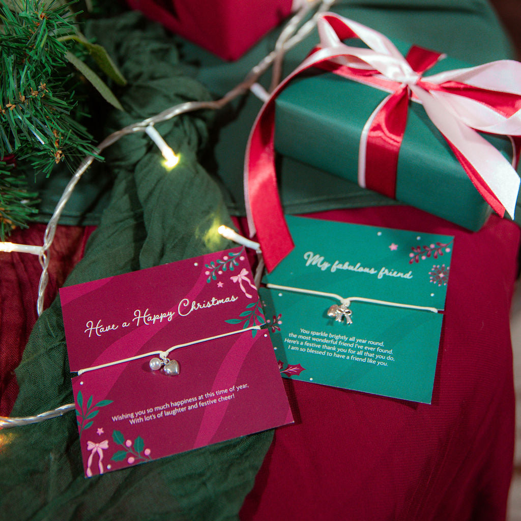 Two wrapped Christmas gift boxes with red and white ribbons and matching gifts from Letterbox Love are arranged on a dark green fabric surface with fairy lights and evergreen branches. The bracelets are elegant gifts for a friend at Christmas.