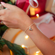 A delicate adjustable bracelet with a small heart & star charm worn on a person's wrist, shown in a festive holiday setting with wrapped gifts and warm string lights in the background.