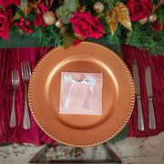 A festive table setting featuring a large metallic bronze charger plate in the center, layered over a deep burgundy tablecloth. A small pink velvet pouch tied with a white ribbon sits on the plate.