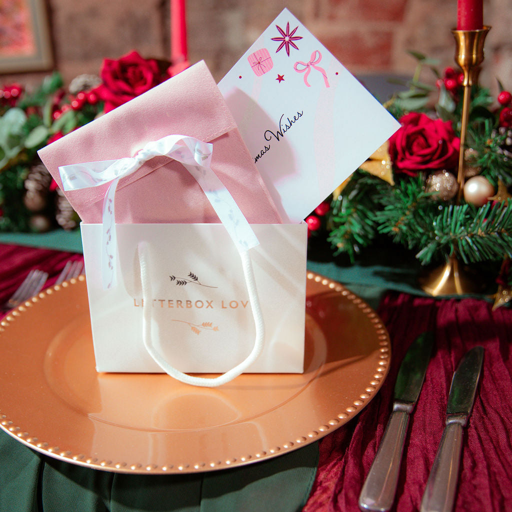 A white and pink gift bag with a white ribbon sits on a gold charger plate, styled with a Letterbox Love postcard on a decorated Christmas table setting.