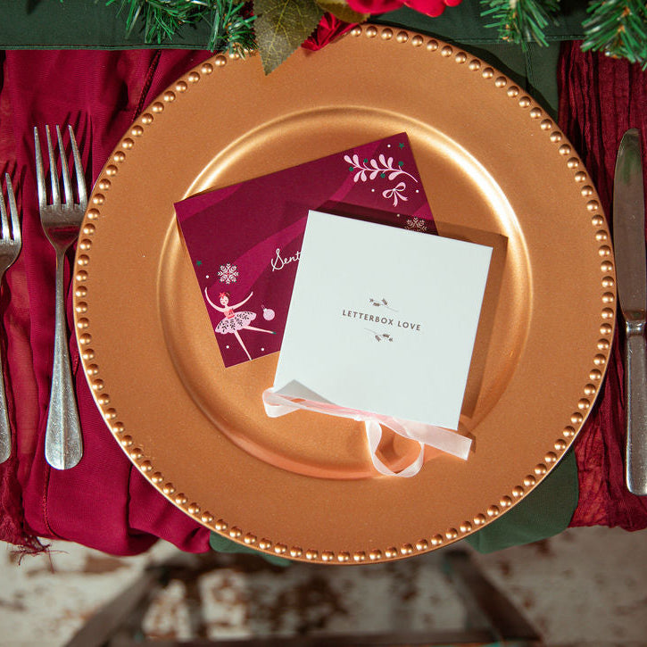 A styled holiday table setting featuring a large gold charger plate, deep burgundy napkins, silver cutlery, and a small white gift box tied with a white ribbon that reads 'Letterbox Love'.