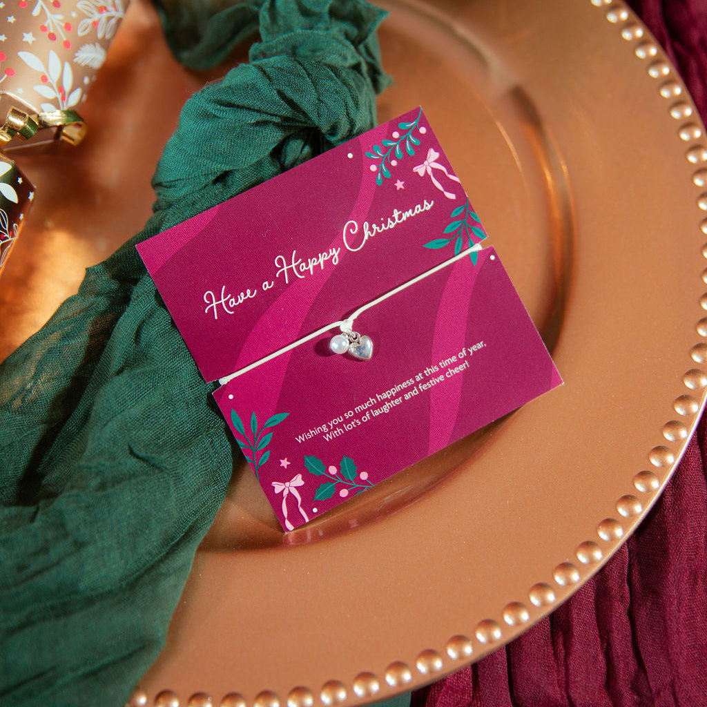 A delicate silver heart bracelet on a matching cord bracelet, presented on a magenta Christmas-themed card, styled on a gold plate with green fabric and holiday decorations.