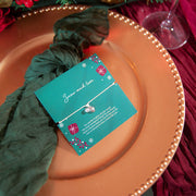 A delicate silver heart bracelet on a green card, styled on a bronze charger plate with a dark green scarf and festive holiday decorations in the background.