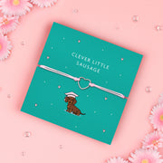A "Clever Little Sausage" bracelet featuring a silver-plated heart charm on a soft pink adjustable cord. The bracelet is displayed on a teal backing card with an illustration of a dachshund wearing a graduation cap. Surrounded by tiny hearts, it's a fun and thoughtful gift for dog lovers and special achievements.