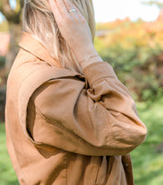 A blonde woman in a brown jacket with her hand touching her hair. On her wrist is a beige adjustable bracelet with a four leaf clover charm.