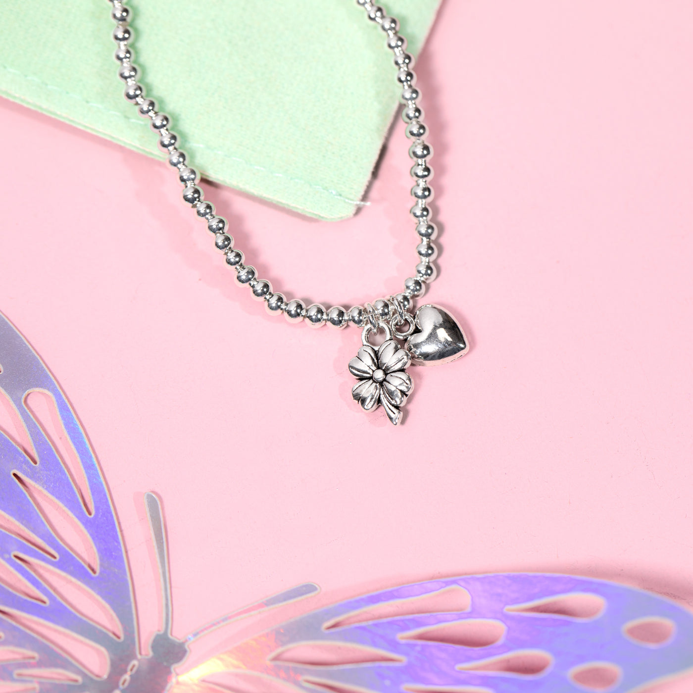 A silver beaded bracelet with a small clover and heart pendant, styled on a pink background with a light green paper corner and decorative butterfly elements.
