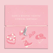 A pink adjustable bracelet featuring a silver-plated cowboy boot charm, displayed on a playful pink backing card with the message "Have a Rootin' Tootin' Cowgirl Birthday" and cowgirl-themed illustrations, including a hat, boots, and cake