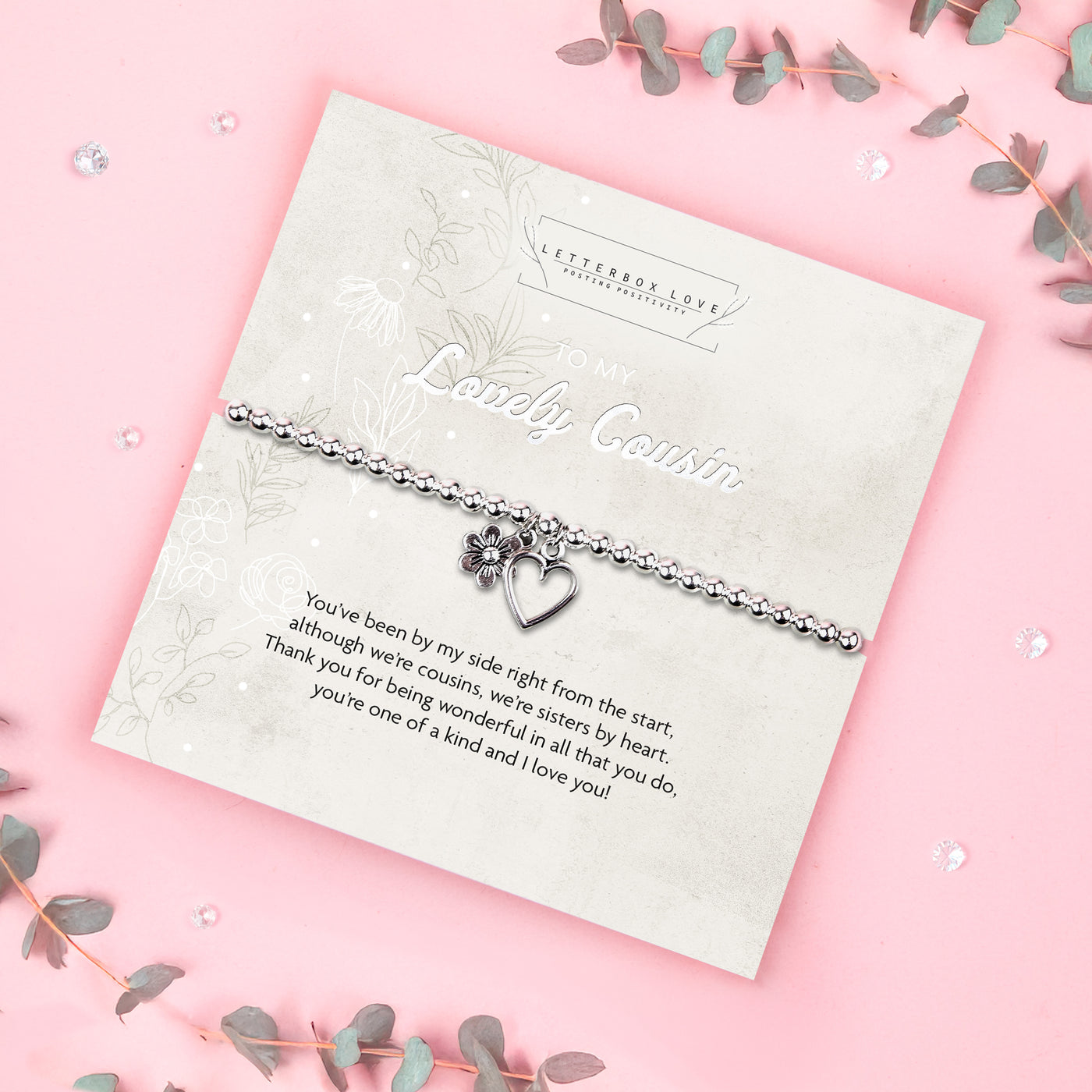 A silver beaded bracelet with a heart and flower charm displayed on a card reading 