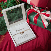 A silver plated beaded bracelet with a small heart charm is displayed inside an open white gift box with a clear lid, placed on red fabric next to wrapped Christmas presents.