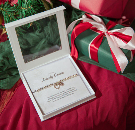 A silver plated beaded bracelet with a small heart charm is displayed inside an open white gift box with a clear lid, placed on red fabric next to wrapped Christmas presents.