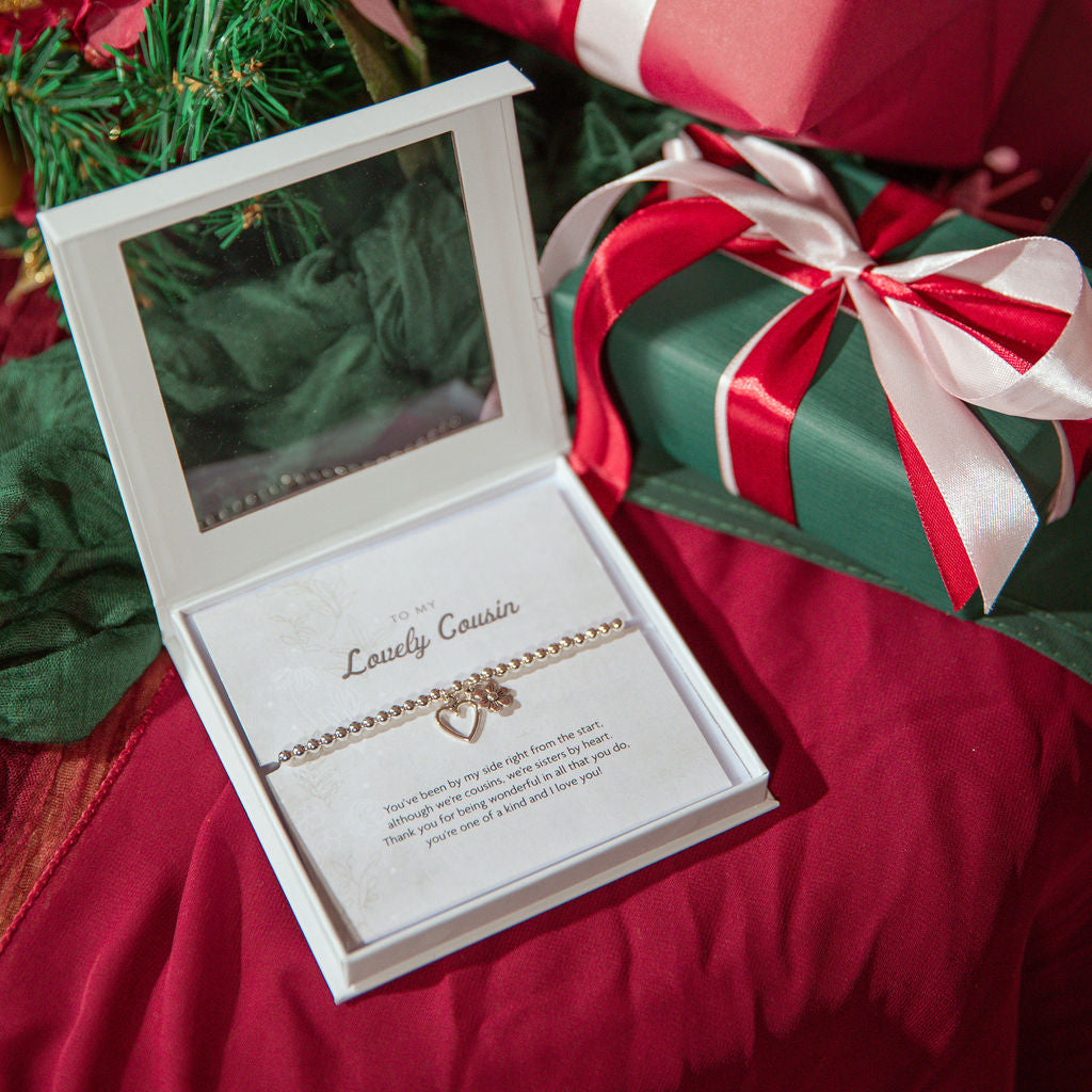 A silver plated beaded bracelet with a small heart charm is displayed inside an open white gift box with a clear lid, placed on red fabric next to wrapped Christmas presents.