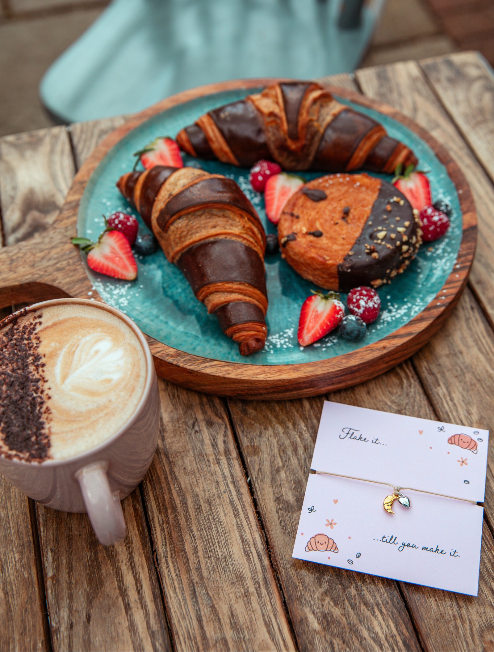 A styled breakfast scene on a wooden table featuring chocolate croissants and a chocolate-dipped pastry on a teal plate with strawberries, alongside a cup of latte and a small card.