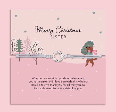 Merry Christmas Sister bracelet featuring a silver wreath charm on an adjustable cord, presented on a festive pink card with snowy trees, stars, and a girl carrying a Christmas tree with her dog. Includes a soft white pouch for gifting and safekeeping. A sentimental Christmas jewellery gift for sisters, perfect as a stocking filler, Christmas morning surprise, or heartfelt keepsake to celebrate sibling love during the holiday season.