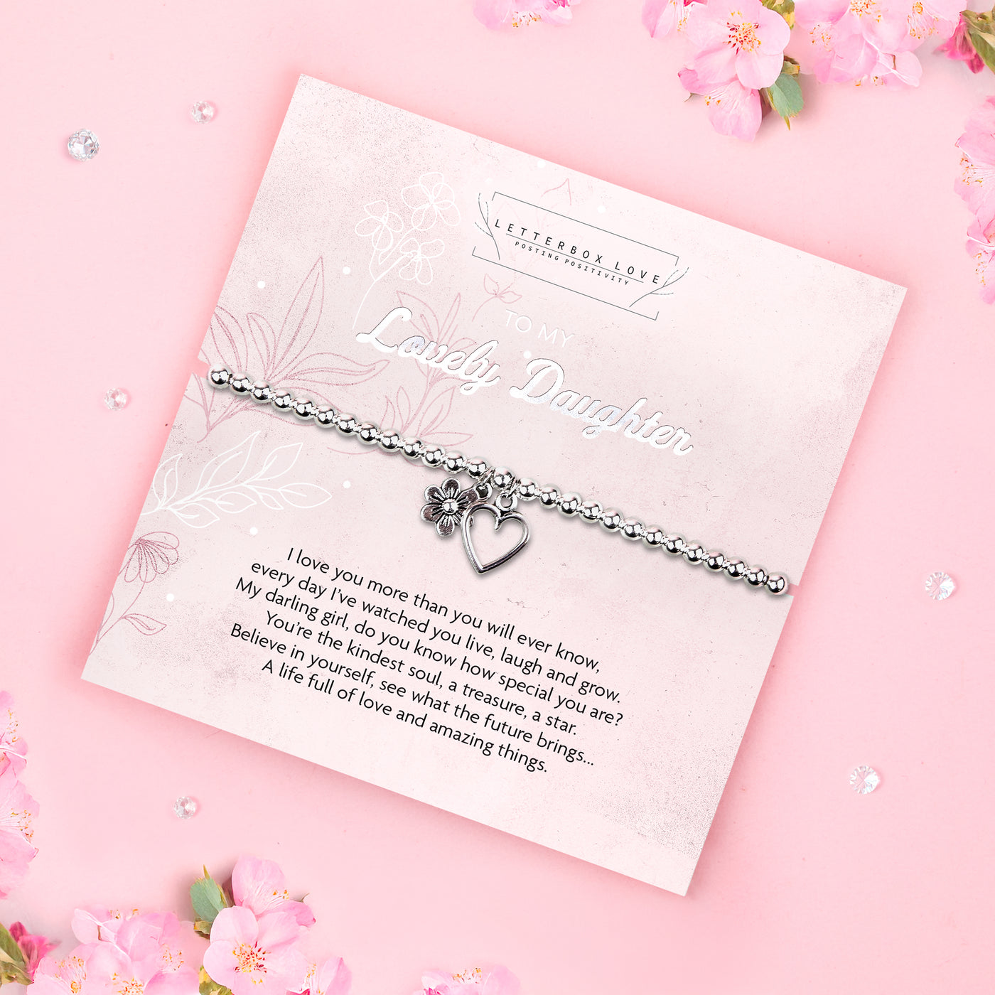 A silver beaded bracelet with a heart and flower charm displayed on a card reading 