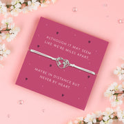 A white adjustable bracelet featuring a heart charm with a world map design, displayed on a deep pink backing card with the message: "Although it may seem like we’re miles apart, maybe in distance but never by heart." Surrounded by white blossoms. Perfect for gifting.