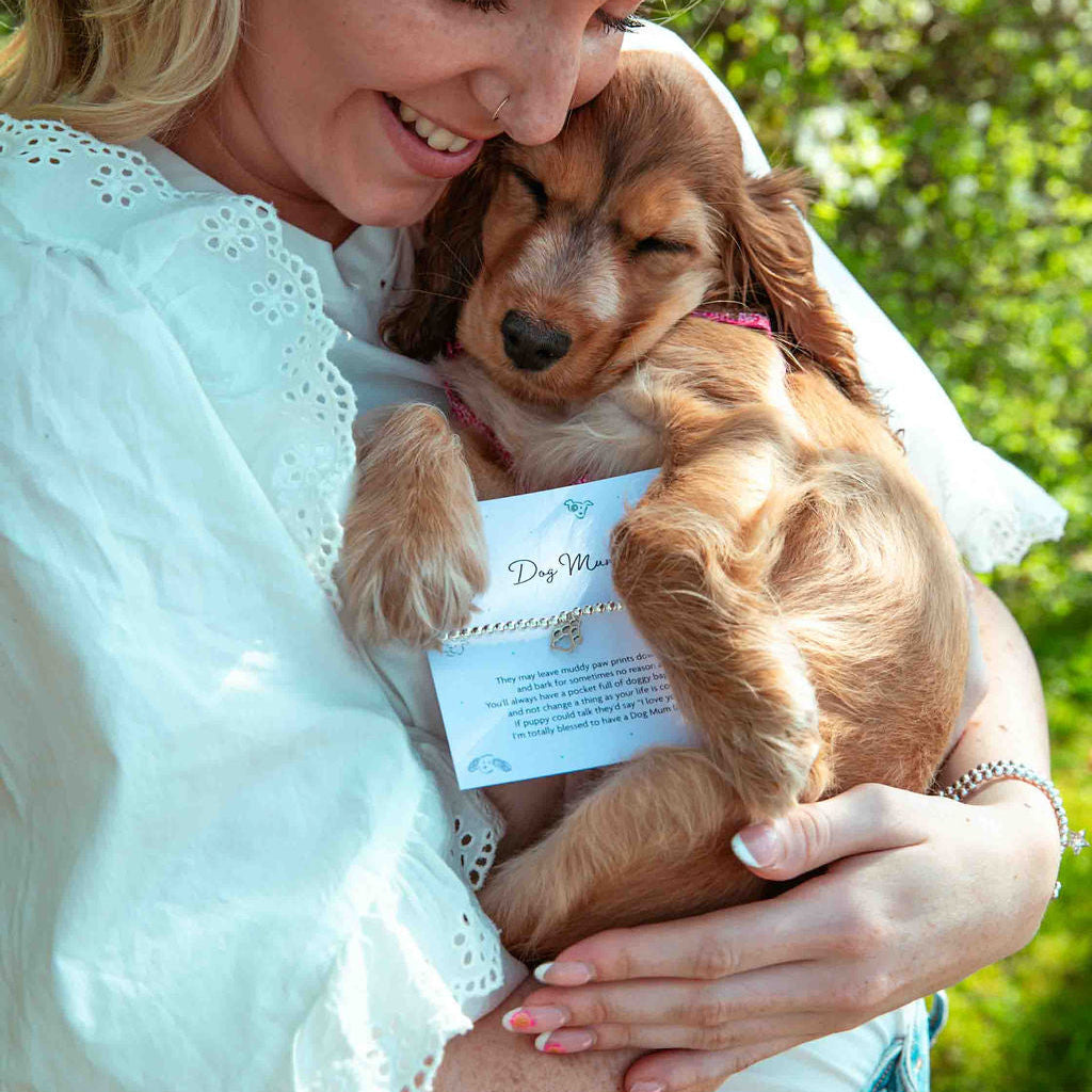 Woman holding a small brown dog with a dog mum poem card with elasticated silver-plated bracelet. A beautiful gift idea for dog lovers. They're standing outside with bushes behind them.