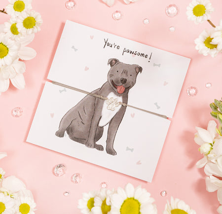 A playful greeting card set on a soft pink background, featuring a hand-drawn illustration of a smiling grey dog with the words 'You're pawsome!' above. The dog is adorned with a shimmering paw charm attached by a thin cord.