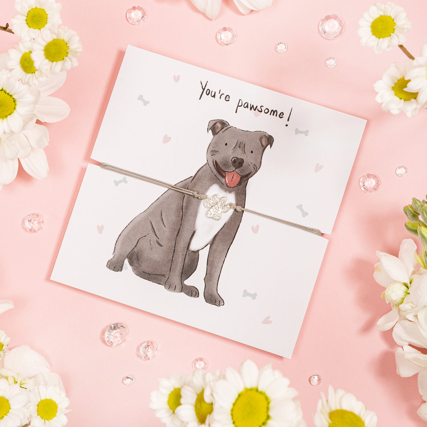 A playful greeting card set on a soft pink background, featuring a hand-drawn illustration of a smiling grey dog with the words 'You're pawsome!' above. The dog is adorned with a shimmering paw charm attached by a thin cord.