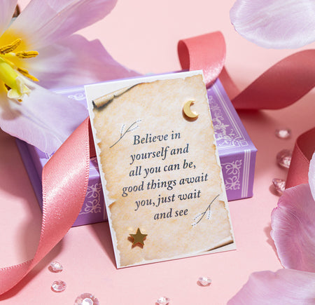 A pair of elegant star-shaped gold earrings resting on a vintage-style paper card within a purple gift box. The card reads 