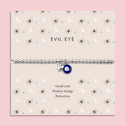Silver-plated beaded bracelet featuring a deep blue evil eye charm and a small heart charm, displayed on a cream sentiment card with moon and star illustrations and the words “Good Luck, Positive Energy, Protection.