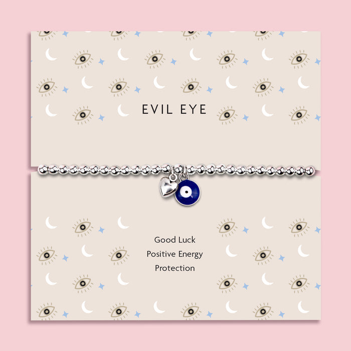 Silver-plated beaded bracelet featuring a deep blue evil eye charm and a small heart charm, displayed on a cream sentiment card with moon and star illustrations and the words “Good Luck, Positive Energy, Protection.