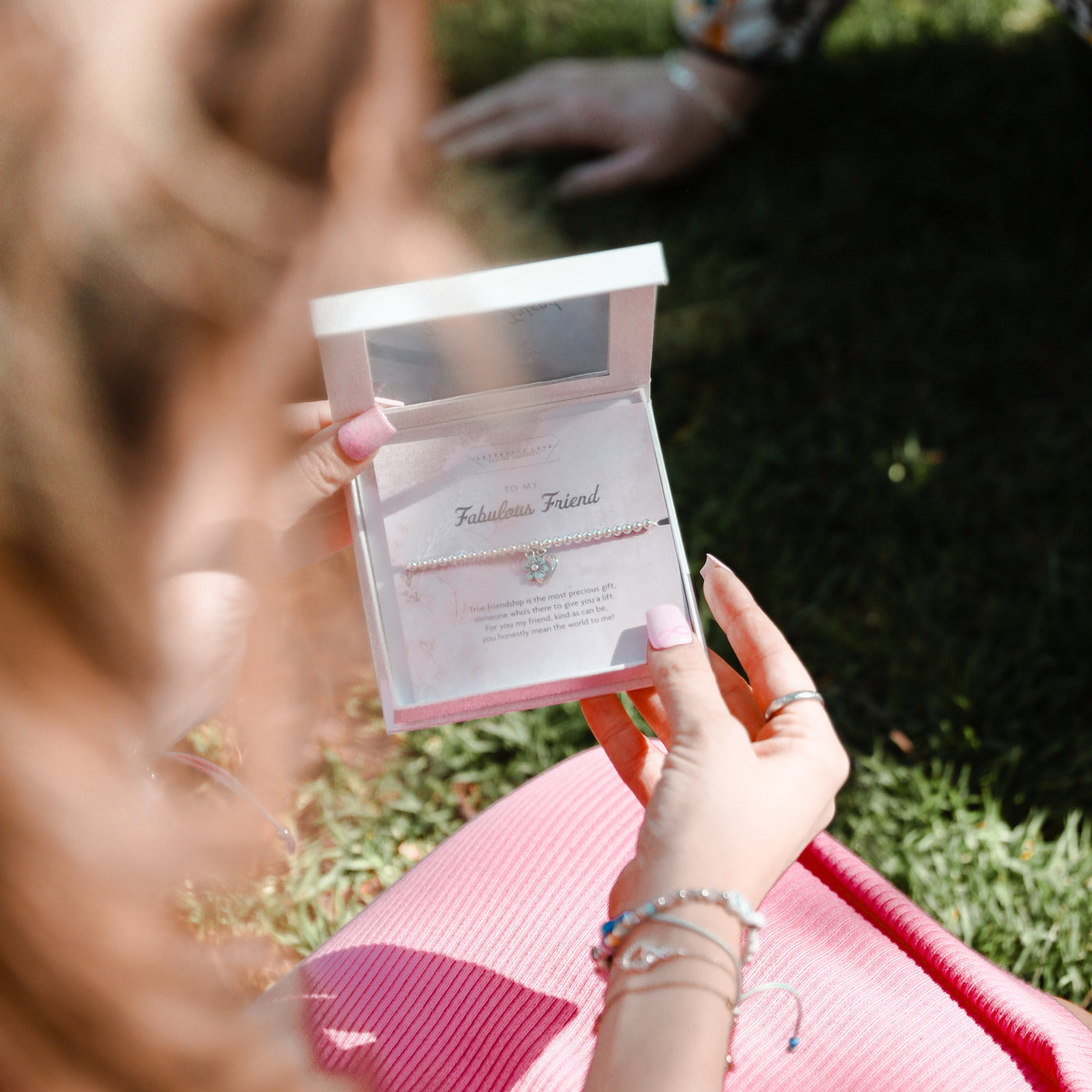 A person outdoors on grass holds an open jewelry box containing a delicate bracelet with a small heart pendant. The person is wearing a pink top and multiple bracelets, with another person partially visible in the background.