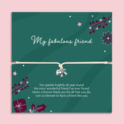 A square green xmas bracelet gift with decorative illustrations and a beige cord across the centre with a silver bow and heart charm. The top half reads 'My fabulous friend' and the bottom half has a festive thank-you message, all on a light pink background.