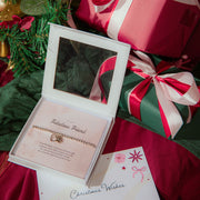 A close-up of a gift box containing silver plated beaded bracelet with a small heart charm, displayed on a pink card that reads 'Fabulous Friend.' The box is surrounded by wrapped Christmas presents and festive greenery, with a card reading 'Christmas Wishes' in the foreground.