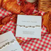 Two delicate silver star-themed friendship bracelets on white cards, styled on a red and white checkered blanket with autumn leaves, a croissant, and coffee cups.