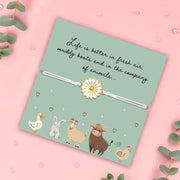 A pale pink background with a green animal poem card with cute illustrations. The bracelet around the card is a white adjustable cord with a white daisy charm. The poem card is surrounded by gems and eucalyptus. 