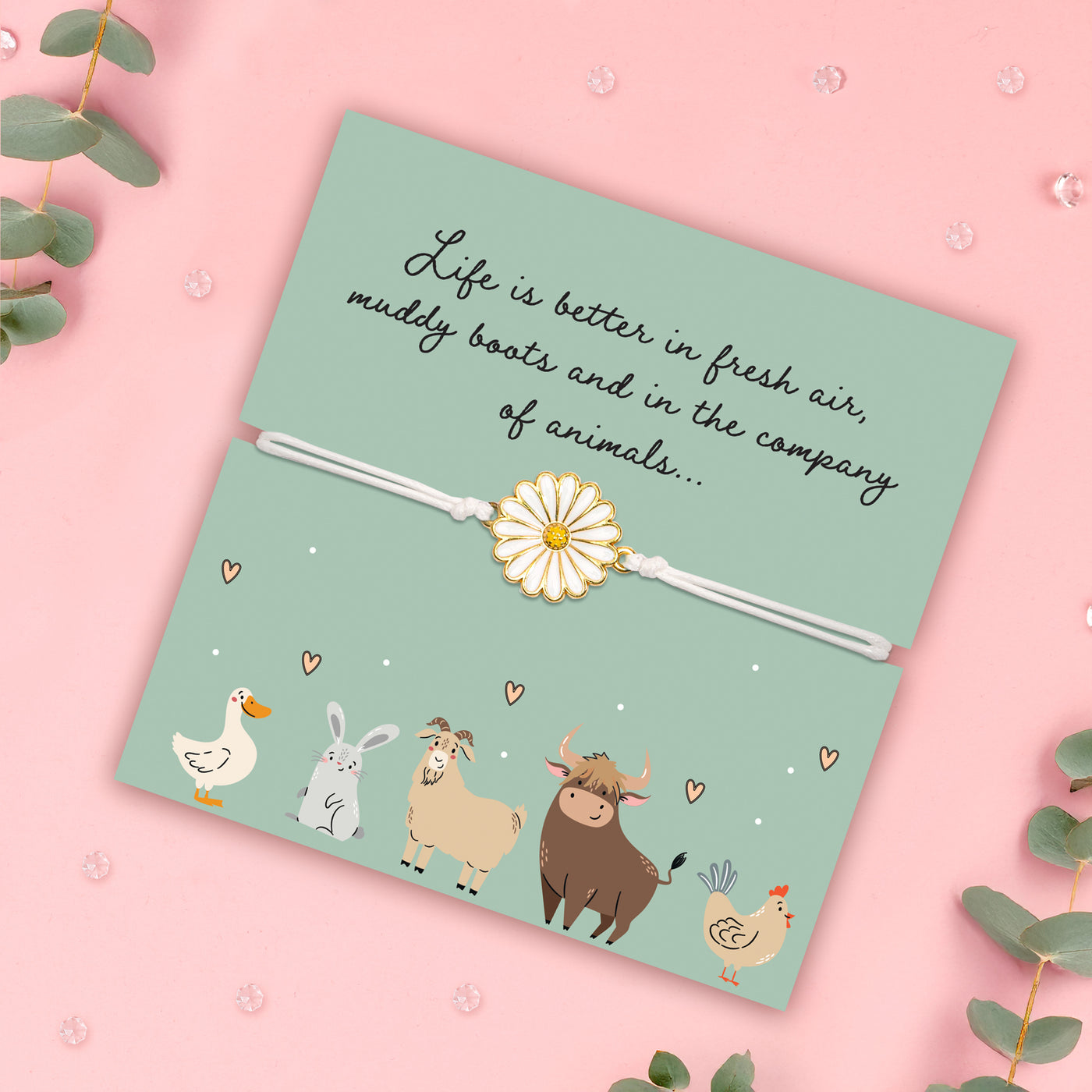 A pale pink background with a green animal poem card with cute illustrations. The bracelet around the card is a white adjustable cord with a white daisy charm. The poem card is surrounded by gems and eucalyptus. 