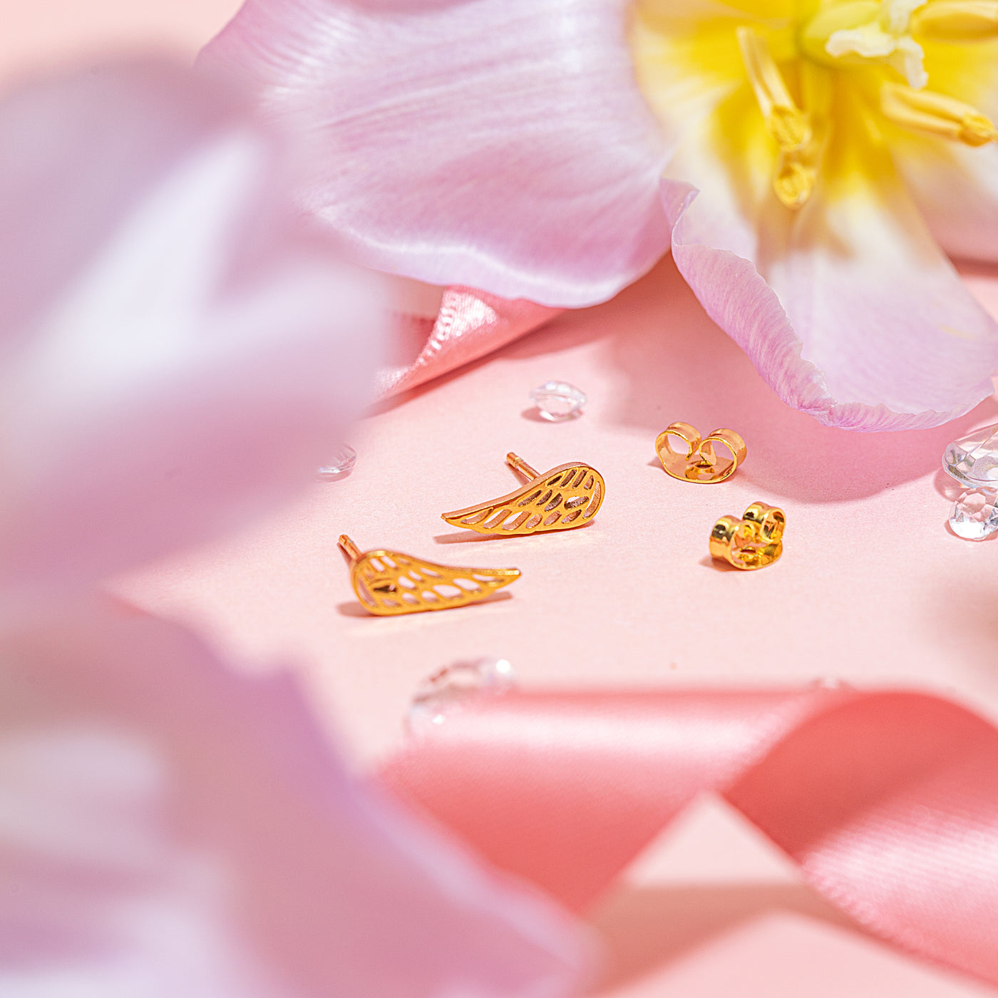 Close-up image of tiny gold feather-shaped stud earrings with butterfly backs, styled on a soft pink background with floral petals and satin ribbon, part of the Letterbox Love earring collection.