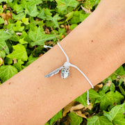 A silver-toned charm bracelet with a feather and heart pendant on a white cord, shown worn on a person's wrist against a background of green ivy leaves.