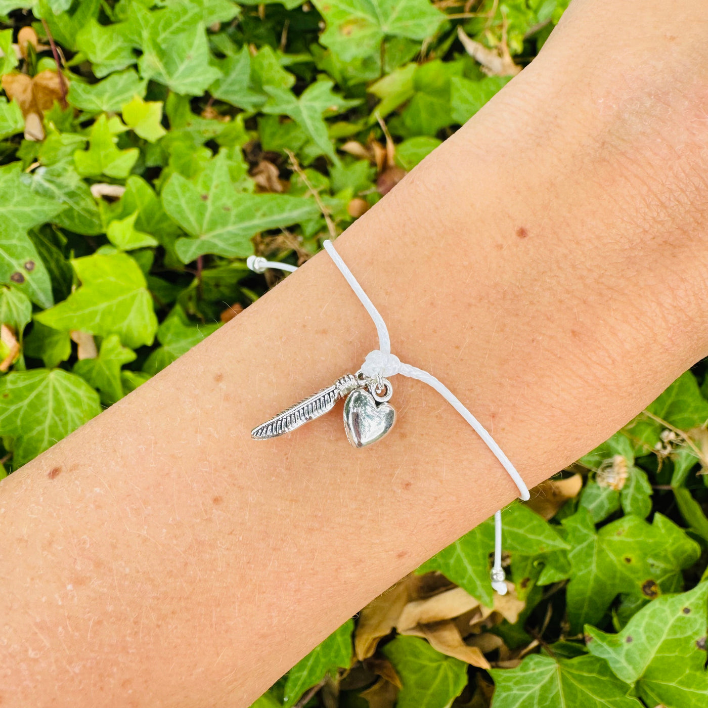 A silver-toned charm bracelet with a feather and heart pendant on a white cord, shown worn on a person's wrist against a background of green ivy leaves.