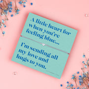 A teal-colored bracelet card titled "Feeling Blue" from Letterbox Love, featuring a small heart charm on a grey string bracelet. The card includes the message: "A little heart for when you’re feeling blue... I’m sending all my love and hugs to you." The setting is a soft pink background with small blue flowers and decorative gems scattered around.