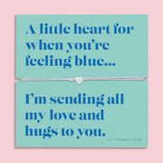 A teal-colored bracelet card titled "Feeling Blue" from Letterbox Love, featuring a small heart charm on a grey string bracelet. The card includes the message: "A little heart for when you’re feeling blue... I’m sending all my love and hugs to you." The setting is a soft pink background with small blue flowers and decorative gems scattered around.